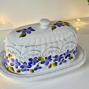 May include: White ceramic butter dish with a lid, decorated with hand-painted purple flowers and green leaves. The dish has a scalloped edge and a small knob on top. A decorative piece for the kitchen.