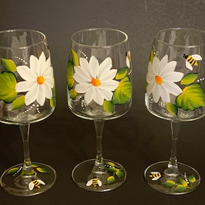 May include: Set of three hand-painted wine glasses with white daisies and green leaves. Each glass features a small yellow and black bee.