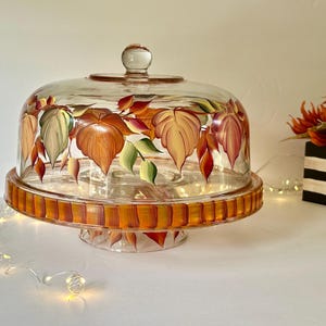 May include: A clear glass cake stand with a matching dome lid, featuring hand-painted autumn leaves in shades of orange, red, and green. The base of the stand is decorated with a series of vertical orange and brown stripes around the rim.