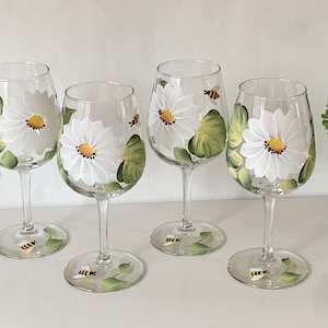 May include: Four clear glass wine glasses with hand-painted white daisies and green leaves. Each glass features a small yellow and black bee.
