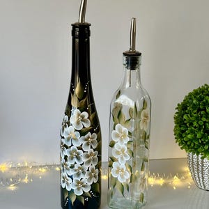 May include: Two glass bottles with hand-painted white flowers on a black and green background. The bottles have silver pour spouts and are perfect for oil and vinegar.