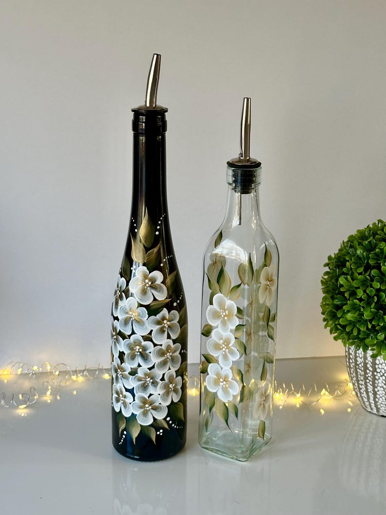 May include: Two glass bottles with hand-painted white flowers on a black and green background. The bottles have silver pour spouts and are perfect for oil and vinegar.