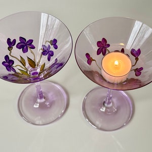 May include: Two purple glass martini glasses with painted purple flowers and green leaves. One glass has a lit tea light candle inside.