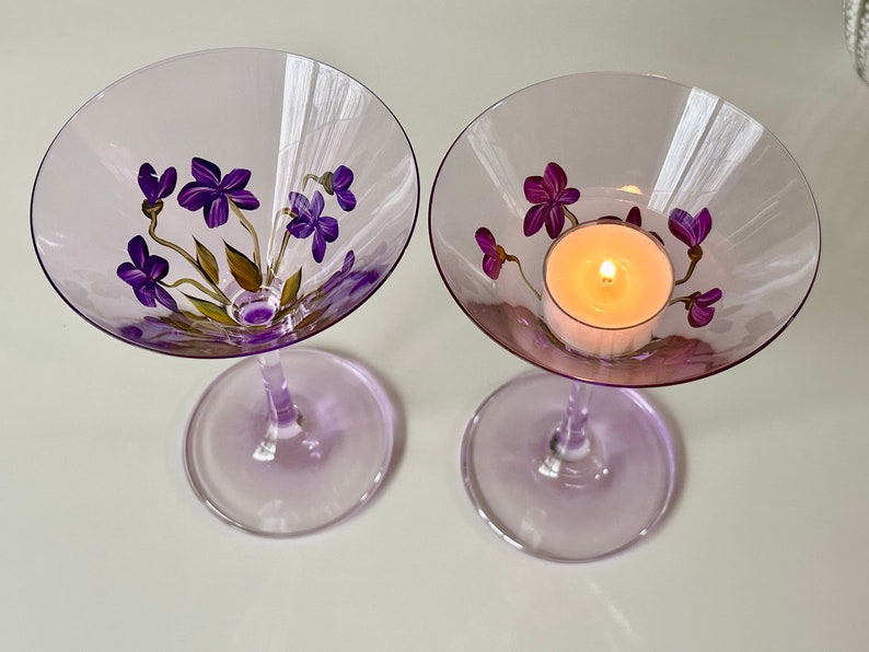 May include: Two purple glass martini glasses with painted purple flowers and green leaves. One glass has a lit tea light candle inside.