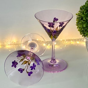 May include: Two purple glass martini glasses with hand-painted purple flowers. The glasses are on a white surface with a string of white lights in the background.