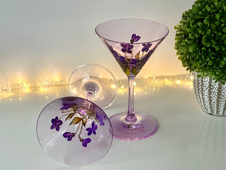 May include: Two purple glass martini glasses with hand-painted purple flowers. The glasses are on a white surface with a string of white lights in the background.
