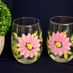 May include: Two clear stemless wine glasses, each hand-painted with a pink daisy design. The flowers have yellow centers and are surrounded by green leaves. The glasses are set against a dark blue background.