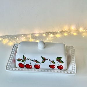 May include: A white ceramic butter dish with a lid and a tray. The lid is adorned with a hand-painted design of red cherries with green leaves. The tray has a beaded edge and a white knob on top.