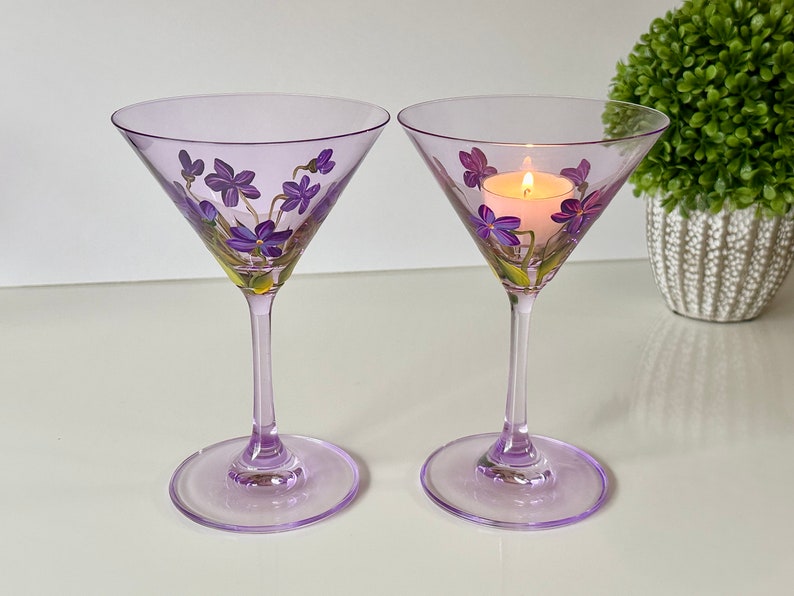 May include: Two purple glass martini glasses with hand-painted purple flowers. One glass has a lit tea light inside.