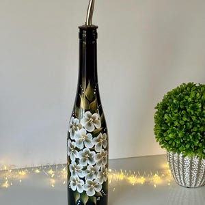 May include: A black glass bottle with a silver pour spout, hand-painted with white and gold flowers. The bottle is decorated with a floral design.