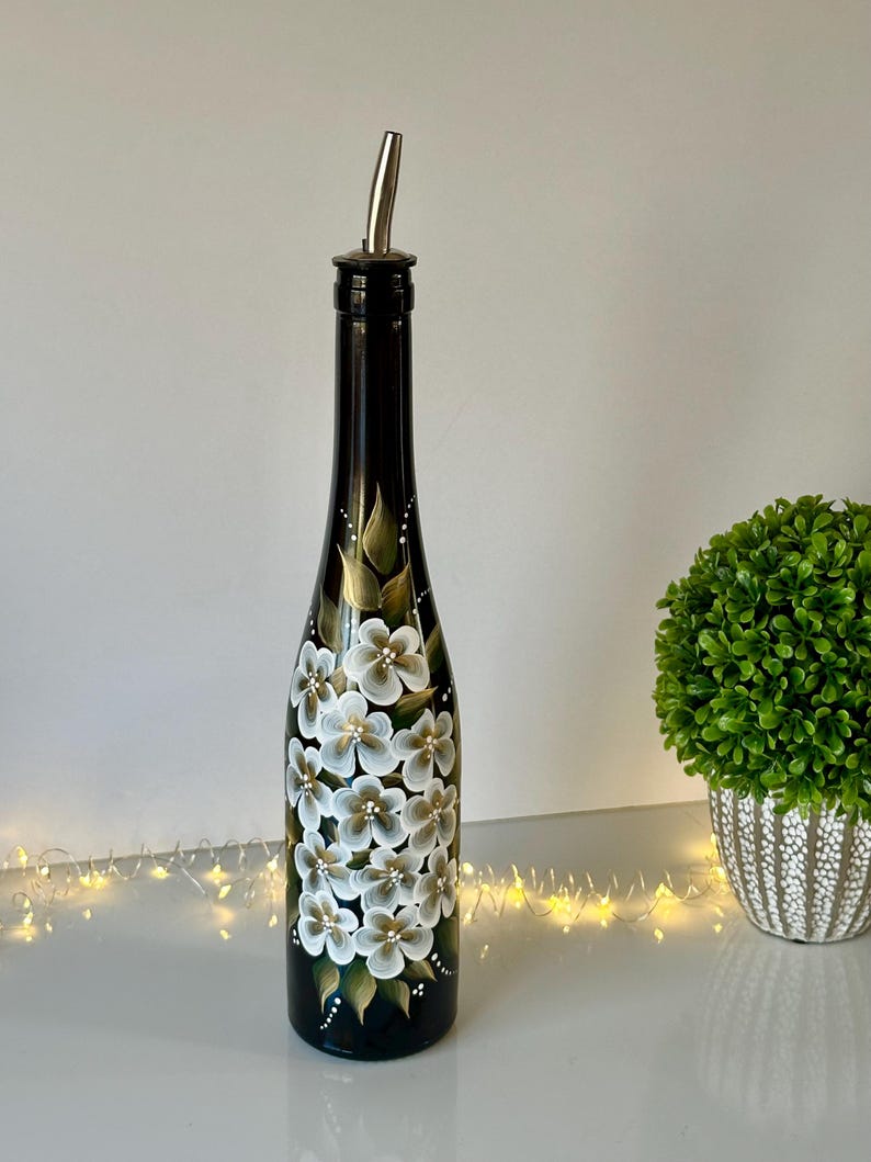 May include: A black glass bottle with a silver pour spout, hand-painted with white and gold flowers. The bottle is decorated with a floral design.