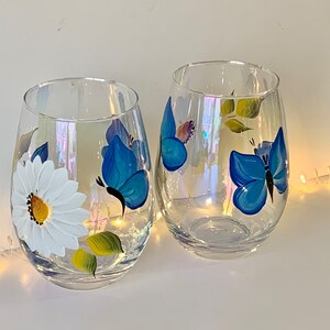 May include: Two clear glass wine glasses with hand-painted designs. One glass features a white daisy with yellow center and green leaves. The other glass has two blue butterflies with black outlines.