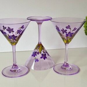 May include: Three purple glass martini glasses with hand-painted purple flowers. The glasses are on a white surface.