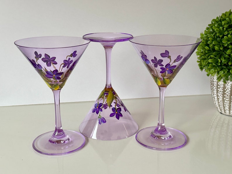 May include: Three purple glass martini glasses with hand-painted purple flowers. The glasses are on a white surface.