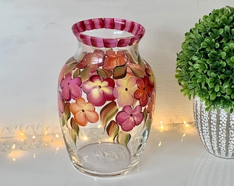 Puede incluir: Un jarrón de vidrio transparente con un borde rayado rosa y blanco. El jarrón está decorado con flores pintadas a mano en rosa, naranja y amarillo.