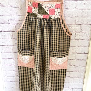 May include: A handmade apron with a patchwork top featuring pink squares and floral patterns. The main body is black and tan plaid with two pink pockets adorned with lace. The apron hangs on a wooden hanger.