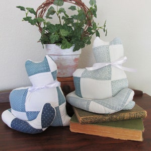 May include: Two handmade cat-shaped pillows. Each pillow is made of patchwork fabric in shades of blue and white, with a white ribbon tied around the neck. The pillows are sitting on a wooden surface.