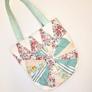 May include: A quilted tote bag with a floral print in shades of pink, yellow, and green. The bag has a light green handle with white polka dots.
