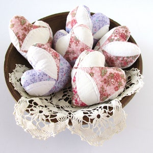 May include: A wooden bowl filled with six handmade fabric hearts. The hearts are made of white fabric with pink and purple floral patterns. The bowl is sitting on a white doily.