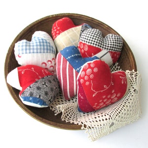 May include: A wooden bowl filled with several heart-shaped fabric ornaments. The hearts are made from various patterned fabrics, including red, blue, and white gingham, stripes, and floral designs. A white lace doily is partially visible.