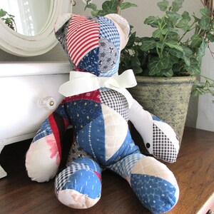 May include: A handmade patchwork teddy bear with a white ribbon around its neck. The bear is made from various fabric patterns, including red and white stripes, blue floral, and black and white gingham. It sits on a wooden surface.