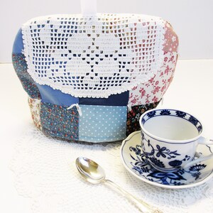 May include: A blue and white patchwork tea cozy with a white crocheted top. The cozy is sitting on a white crocheted doily with a blue and white teacup and saucer.