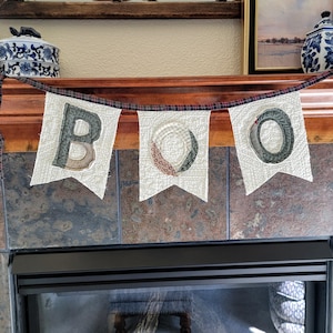 May include: A fabric banner featuring three white quilted pennants suspended from a dark plaid ribbon. The pennants display the letters B, O, and O in dark green and patterned fabric appliqués, creating a decorative hanging word display.