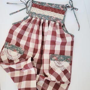 May include: A handmade red and white checkered romper with floral and lace accents. The top features a patchwork design with floral fabric and tie straps. Two pockets with lace and floral trim are on the front.