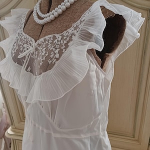 Może przedstawiać: Biała koszula nocna w stylu vintage na manekinie. Koszula nocna posiada koronkowy gorset z delikatną kokardą, falbaniaste rękawy i zwiewną spódnicę. Podwójny sznur sztucznych pereł zdobi szyję manekina.