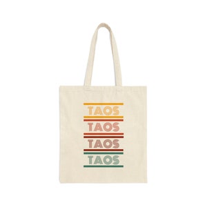 May include: A beige tote bag with the word "TAOS" printed in a retro style in four rows. Each row is a different color: yellow, orange, red, and green.