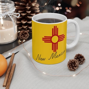 May include: A white ceramic mug with a yellow background featuring the New Mexico Zia symbol in red and a yellow center. The words "New Mexico!" are written in black script. The mug is filled with a dark liquid.