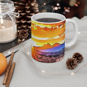 May include: White ceramic mug with a colorful sunset landscape design. The mug is filled with a dark liquid and steam is rising from the top.