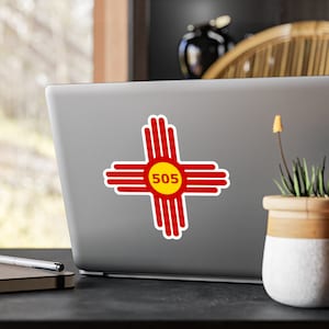 Può includere: Un adesivo rosso e giallo con il simbolo dello stato del Nuovo Messico e il prefisso 505 su un laptop argentato.