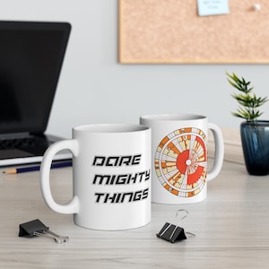 Op de afbeelding: Twee witte keramische mokken. Eén mok heeft de tekst "DARE MIGHTY THINGS" in het zwart gedrukt. De andere mok heeft een kleurrijk cirkelvormig ontwerp met cijfers en letters.