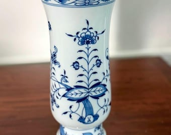 Pode incluir: Um vaso de porcelana branca com um padrão floral azul. O vaso tem um topo alargado, um corpo cilíndrico e uma base de pedestal. O design azul inclui flores, folhas e bordas decorativas. O vaso tem aproximadamente 20 cm de altura.