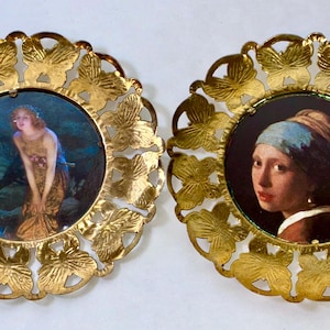 May include: Two circular wall plaques featuring gold-toned metal frames with a repeating butterfly cutout pattern. The left plaque displays a painting of a figure in a landscape, while the right plaque shows a portrait of a person wearing a headscarf.