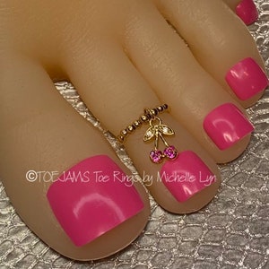 May include: A gold-colored toe ring with a cherry charm featuring pink gemstones. The ring is adorned with small beads and a leaf design. The toenails are painted a bright pink color. The text "TOE JAMS Toe Rings by Michelle Lyn" is visible.