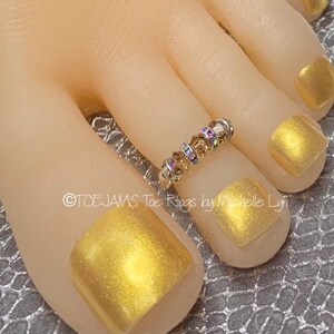May include: A silver toe ring adorned with iridescent and amber-colored crystals. The ring is on a foot with gold nail polish. The text "TOE JAMS Toe Rings by Michelle Lyn" is visible.