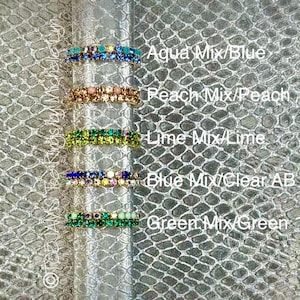 May include: An assortment of colorful rhinestone rings in various shades of blue, peach, lime, and green. Each ring is displayed with its color name: Aqua Mix/Blue, Peach Mix/Peach, Lime Mix/Lime, Blue Mix/Clear AB, and Green Mix/Green. The rings are set against a textured background.