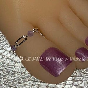 May include: A silver-toned toe ring featuring a delicate chain design with small, round, light purple beads interspersed along the band. The ring is worn on the second toe, which has metallic purple nail polish, and the text TOEJAMS Toe Rings by Michelle Lyn is visible.