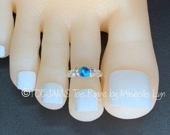 May include: A toe ring featuring a central bright blue marbled bead flanked by smaller clear, pink, and purple accent beads on a metallic band. The text TOE JAMS Toe Rings by Michelle Lyn is visible across the lower portion of the image.