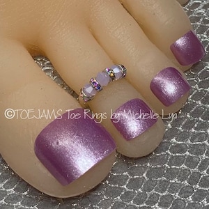 May include: A close-up of a toe ring with light purple beads and iridescent accents. The ring is worn on a toe with purple nail polish. The text "TOEJAMS Toe Rings by Michelle Lyn" is visible.