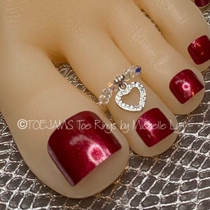 May include: A silver-toned toe ring featuring a dangling heart-shaped charm encrusted with small clear stones. The band is composed of alternating clear faceted beads and small silver-toned spacers. The text TOE JAMS Toe Rings by Michelle Lyn is visible.