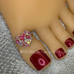 May include: A silver-toned toe ring featuring a heart-shaped centerpiece encrusted with various shades of pink and red rhinestones. The text "TOEJAMS Toe Rings by Michelle Lyn" is printed across the image. The ring is worn on a toe.