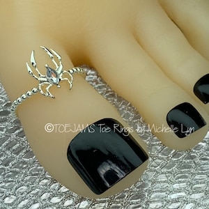 May include: A silver-colored toe ring with a spider design, featuring a central gemstone. The ring is worn on a foot with black nail polish. The text "TOEJAMS Toe Rings by Michelle Lyn" is visible.