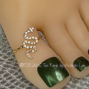May include: A gold-toned toe ring in the shape of a snake, encrusted with small, clear gemstones. The snake design wraps around the toe. The image includes the text "©TOEJAMS Toe Rings by Michelle Lyn". The toenails are painted a dark green.