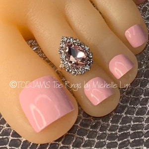 May include: A pink toe ring with a teardrop-shaped rhinestone. The ring is on a model's toe with pink nail polish.