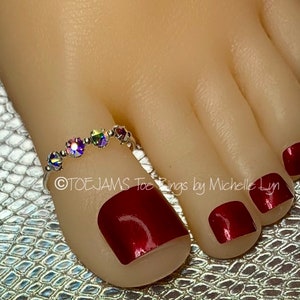 May include: A silver-toned toe ring featuring four small, iridescent, multi-colored circular crystals set in a row. The ring is worn on a toe with dark red nail polish. The text "©TOEJAMS Toe Rings by Michelle Lyn" is visible on the image.