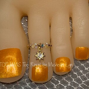 May include: Gold toe ring with a small star charm and clear crystals. The ring is on a foot with gold nail polish.