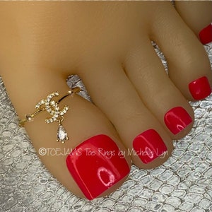 May include: A gold-colored toe ring with a double-C design, adorned with small clear crystals and a dangling teardrop-shaped gem. The toenails are painted a vibrant red. The text "@TOEJAMS Toe Rings by Michelle Lyn" is visible.